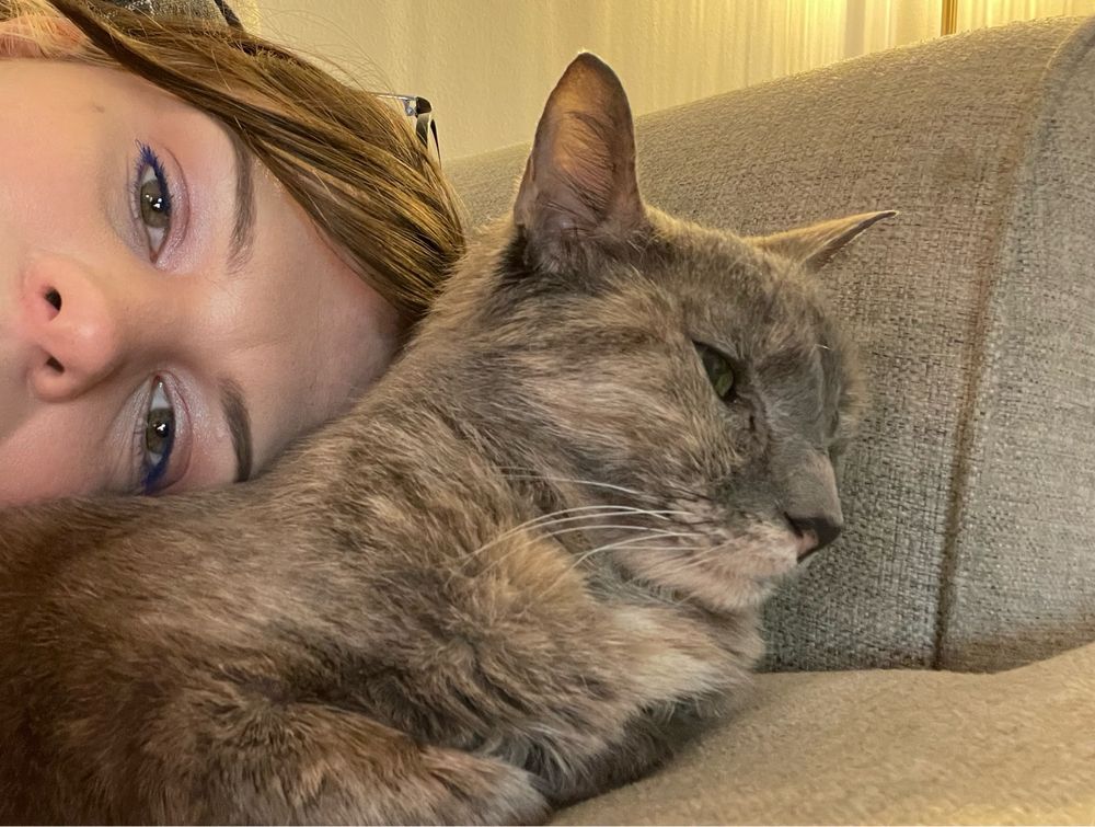 femme presenting person, eyes open, lays her head gently on a gray tortoiseshell cat’s back while the cat stares into the distances 
