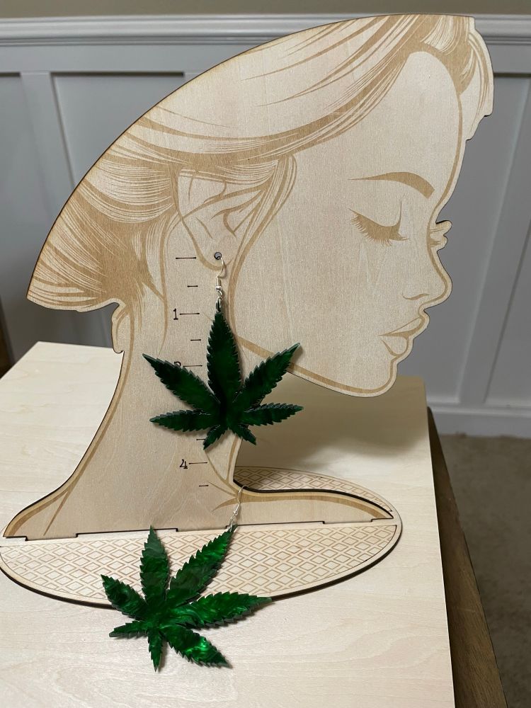 A wooden, laser cutter earring display stand in the shape of a person’s profile, showing a pair of cannabis leaf shaped earrings