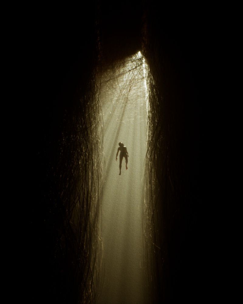 A woman floats between two walls covered in cables, with light shafts coming from above, piercing through a thin fog and a web of cables.
