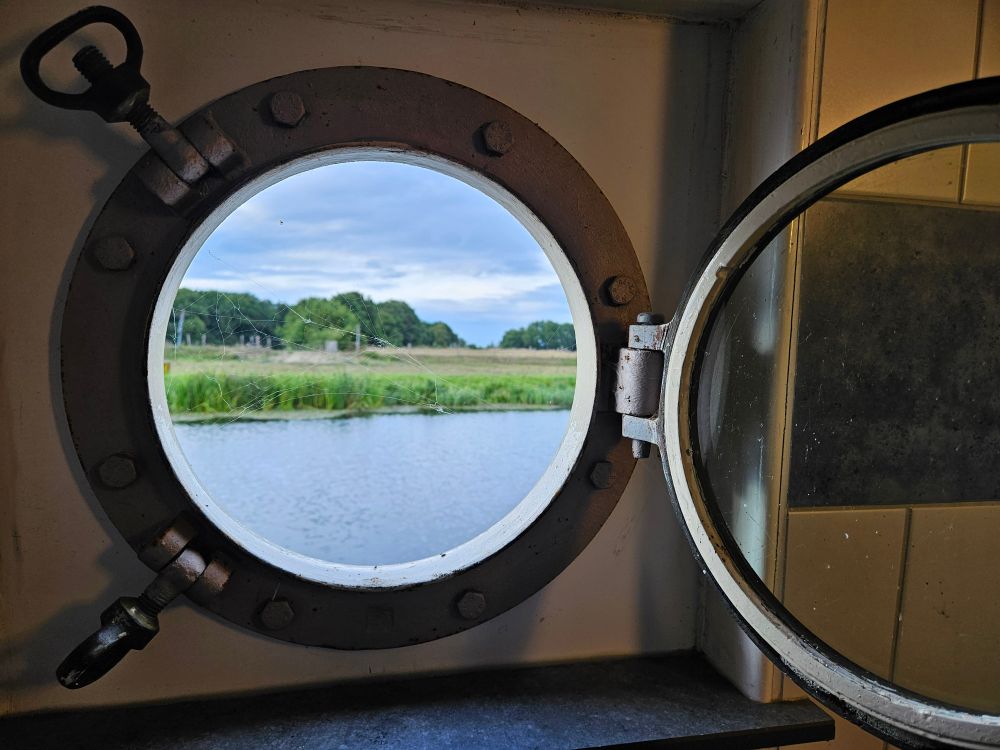 Blik door een opengezet patrijspoort op de oever van de Vecht