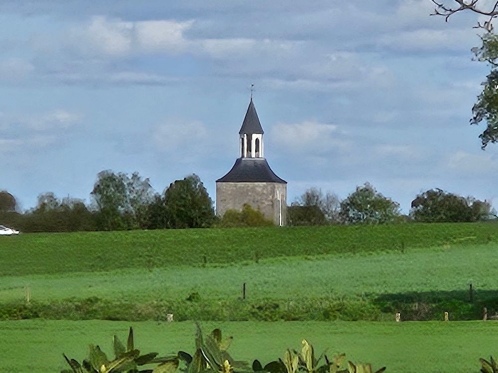 Toren met een brede basis, die sierlijk vernauwt tot een smalle lantaarn met een spitsje erop, samen met een bomenrij achter een grasgroene heuvel onder een halfbewolkte hemel