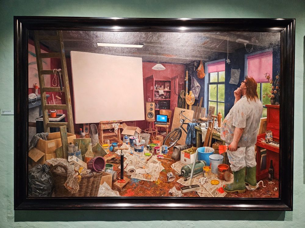 Schilderij "Wachten op inspiratie". Een schilder staat in zijn atelier, chaotisch vol met spullen, voor een leeg doek.