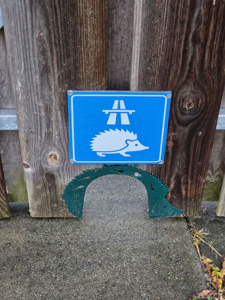 Klein poortje onderin tuinhek met erboven een blauw vierkant bordje met een egel en een autobahnsymbool