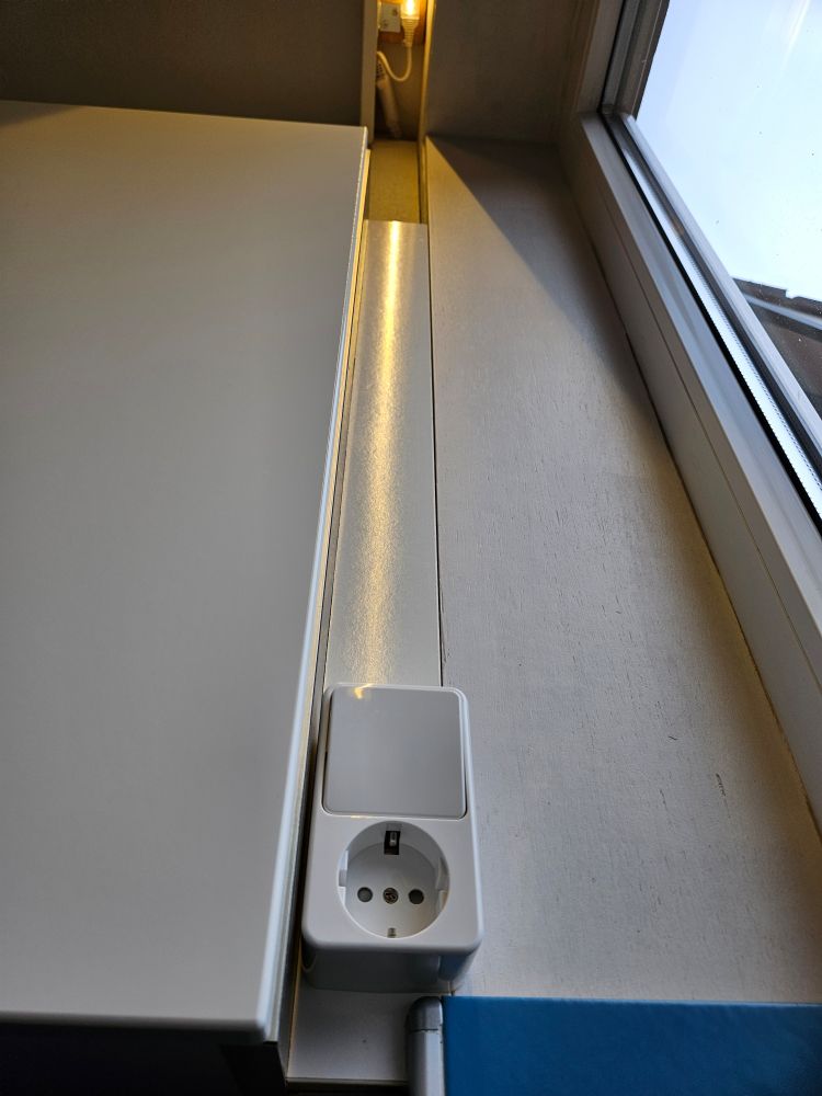 Schakelaar-stopcintactcombinatie op houtwerk tussen keukenkast en raamvenster. De blik van de camera is omhoog, waar de ledstrip boven het raam brandt, weerspiegeld op het glimmende hout.