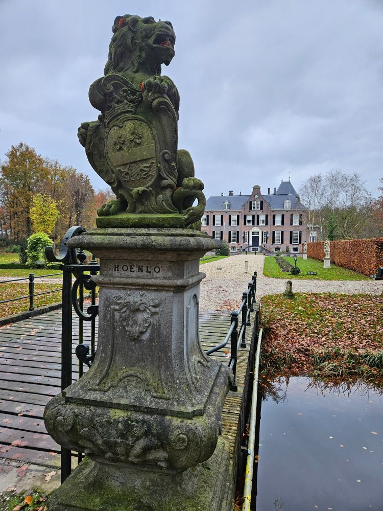 Stenen leeuw met tekst 'Hoenlo' naast toegangsbrug voor het kasteel dat op de achtergrond te zien is.