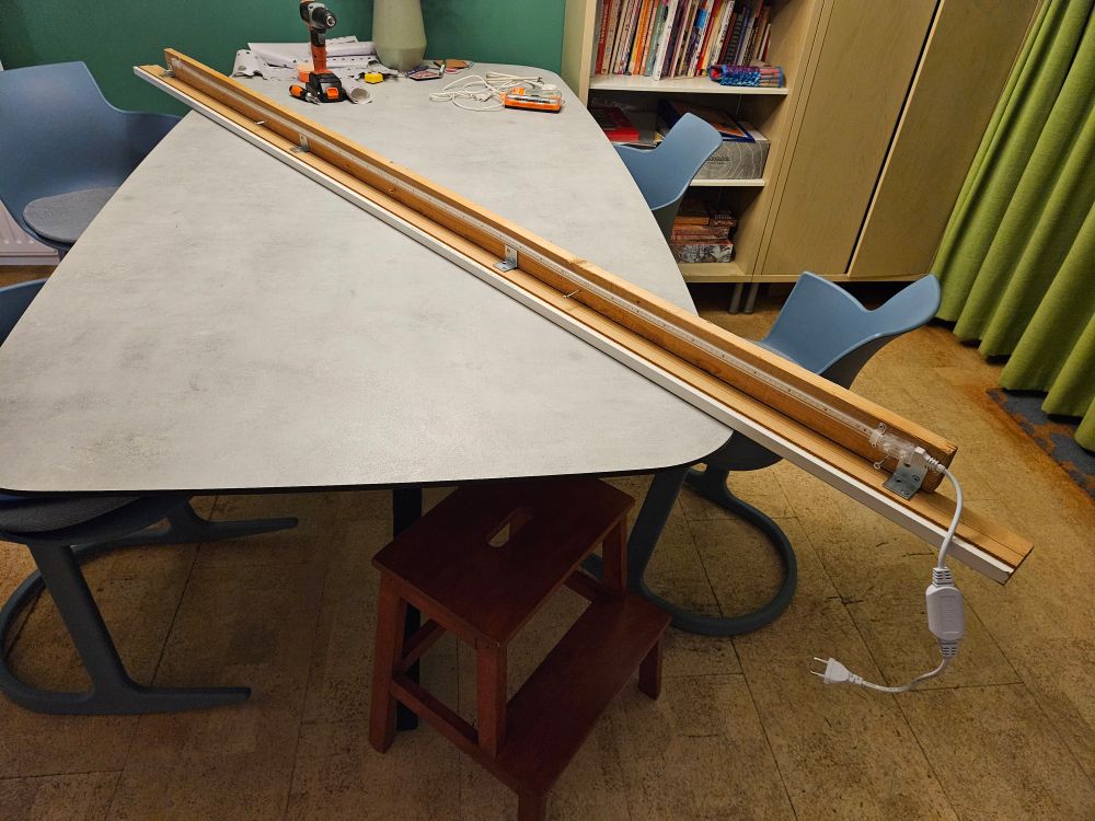 Ledstrip van 2 meter aan een lat waaraan een plint is vastgemaakt met hoekijzertjes, diagonaal liggend op een grijze eettafel. Een stekker bungelt vooraan omlaag.