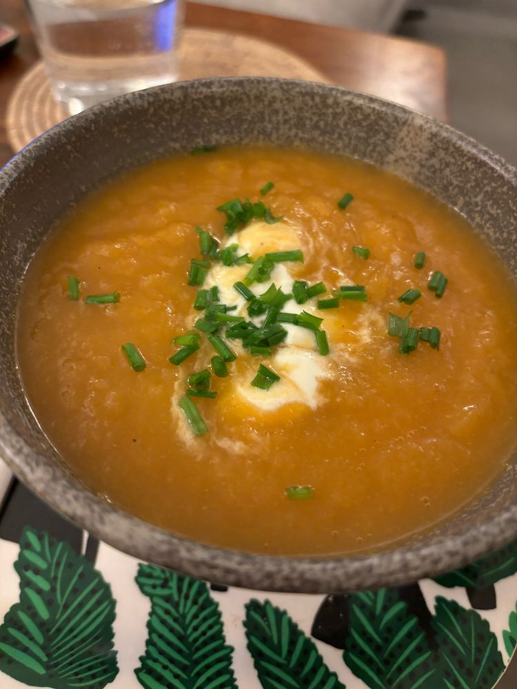 Grey bowl on patterned tray, filled with appetising looking rust coloured soup, topped with yoghurt and cut chives