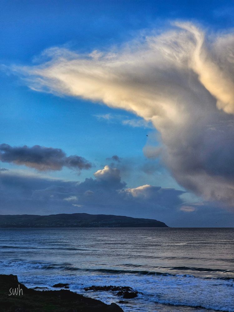 A rocky foreground, then the sea with land in the distance.
Above is some blue sky with dark clouds in the distance and a closer cloud which is catching the sunlight.