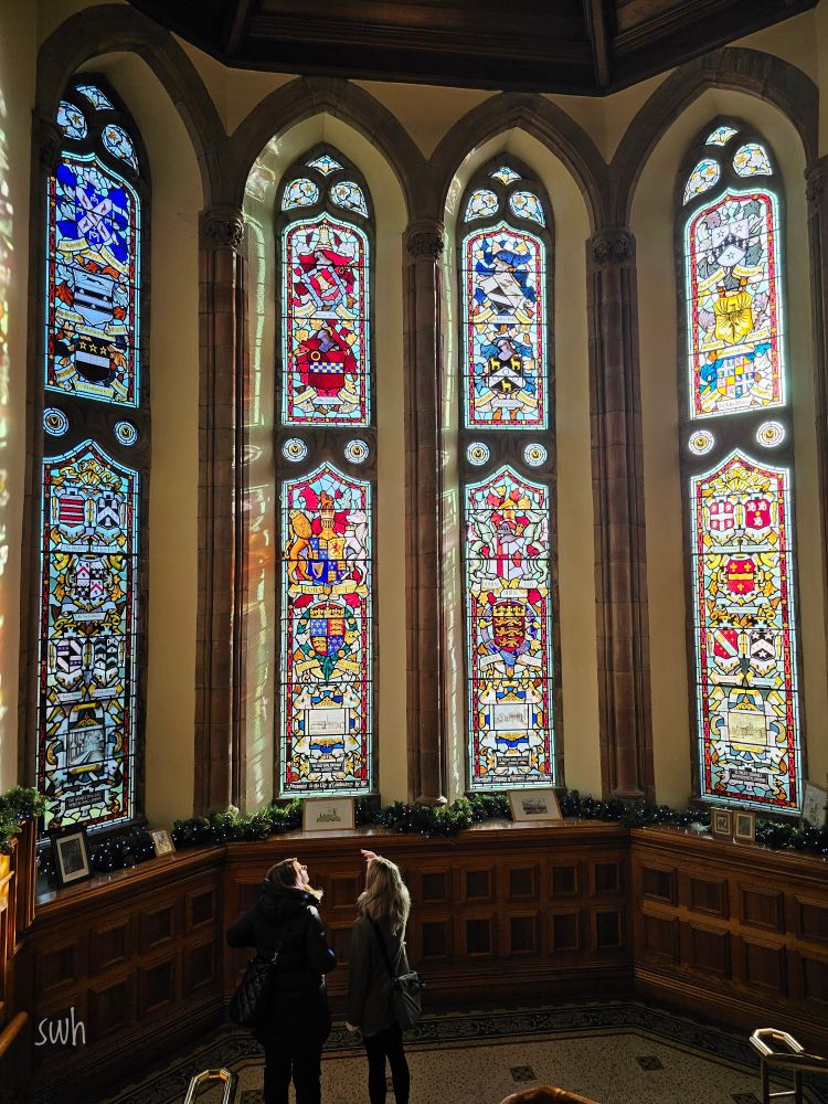Four tall arched stained glass windows, with two people looking at the windows.
Each window has a very intricate design incorporating various coats of arms.
