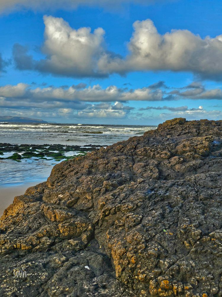 Looking past a large rock towards the sea.
With blue sky and fluffy clouds.