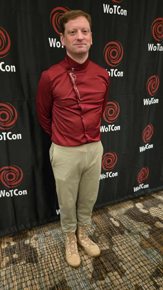 A man stands with feet close together, hands behind his back, in a red sateen shirt with buttons running diagonally up the front. He has a silver lion pin at the top of the buttons at his throat. His shirt is tucked into loose-fitting tan trousers, which are tucked into tan boots. 