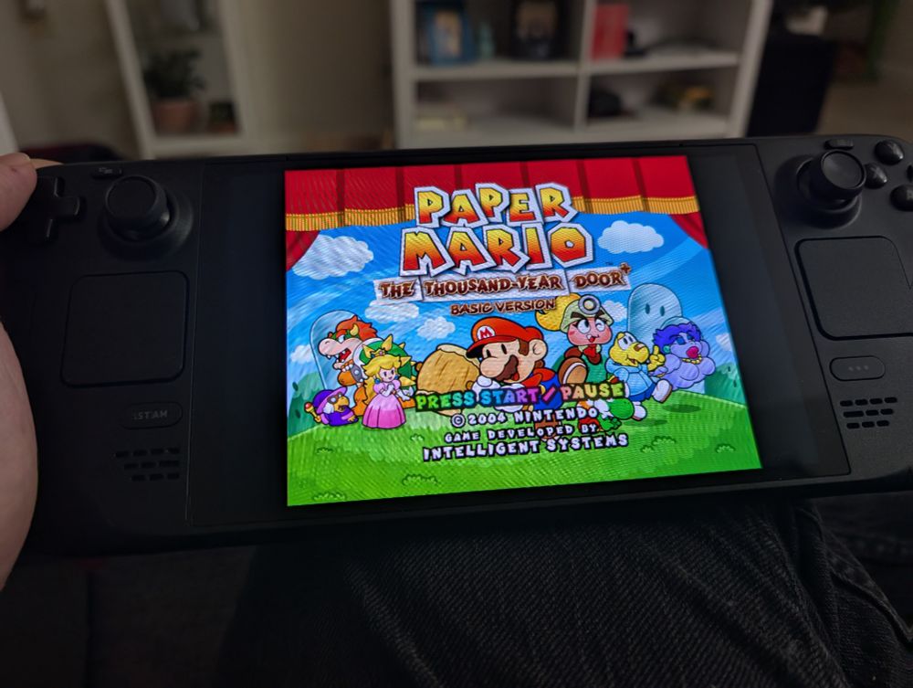 A handheld gaming device, a Steam Deck, displays the title screen of the video game "Paper Mario: The Thousand-Year Door." The game's vibrant, paper-thin characters are shown on a stage-like background with red curtains. The screen text reads "PAPER MARIO THE THOUSAND-YEAR DOOR" and below that "BASIC VERSION." Instructions "PRESS START PAUSE" are also visible.
