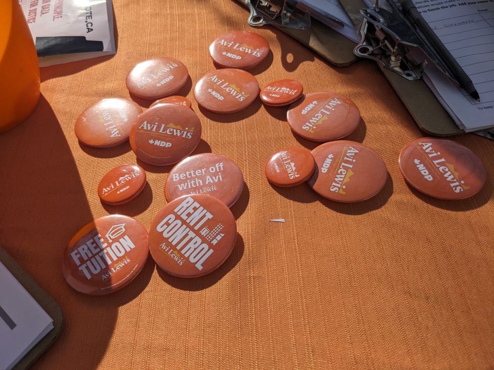 A sunwashed table covered with orange NDP badges relevant to the Vancouver candidate Avi Lewis.