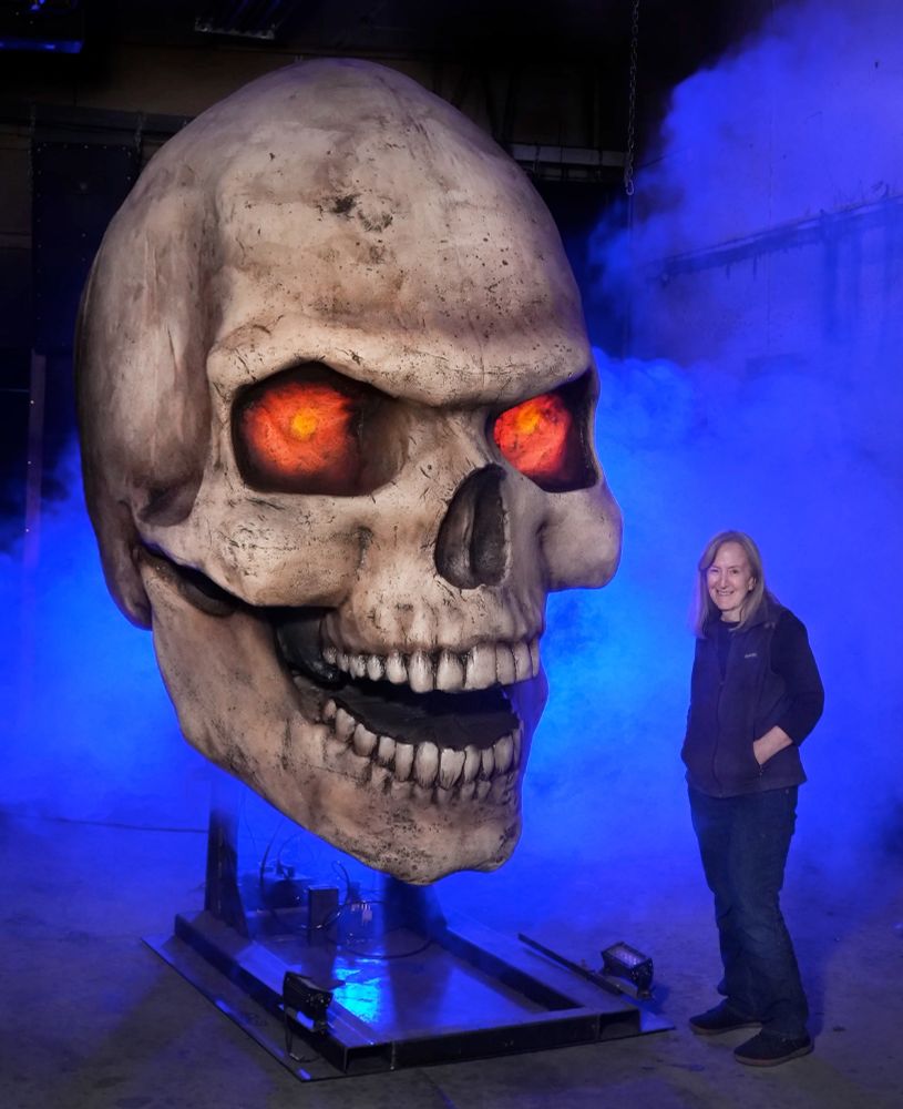 A smiling woman standing next to a truly gigantic animatronic prop of a skull with glowing red eyes