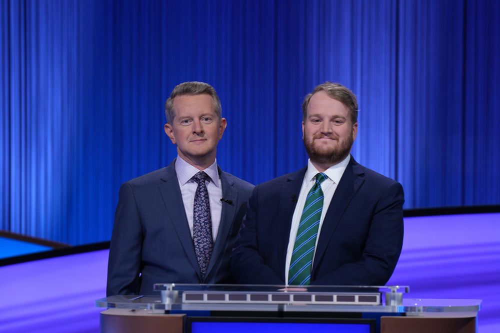 Ken Jennings and contestant Matt Massie stand behind a podium on the Jeopardy stage and smile for the camera