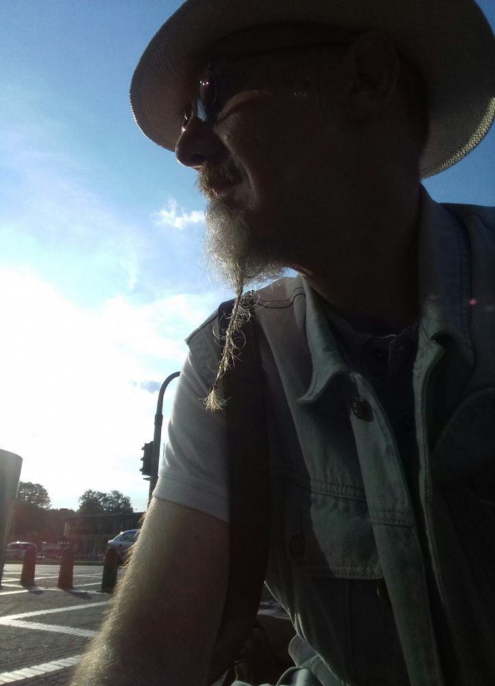 Alter Mann auf einer nicht sichtbaren Sitzgelegenheit. Der Himmel ist blau, Wolken sind zu sehen. Er trägt einen Panamahut und eine Jeansweste. Sein Bart ist geflochten.