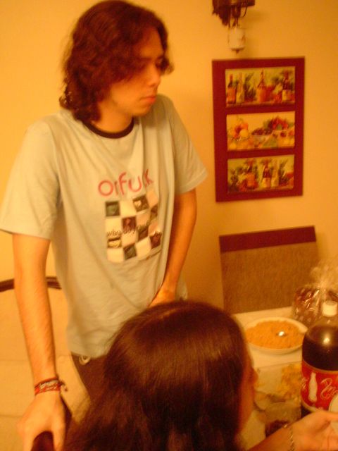 foto minha de 2005, estou de pé, apoiado em um encosto de cadeira, vestindo uma blusa azul, sem barba e com cabelos ondulados de tamanho médio