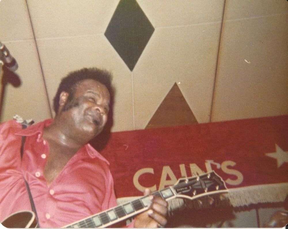 Freddie King playing electric guitar, “Cain’s is printed or painted on the wall behind him