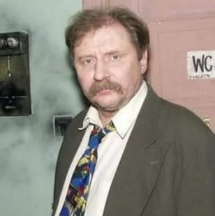 Ferdynand Kiepski w koszuli i pod krawatem, stoi pod publiczno-prywatną toaletą na korytarzu