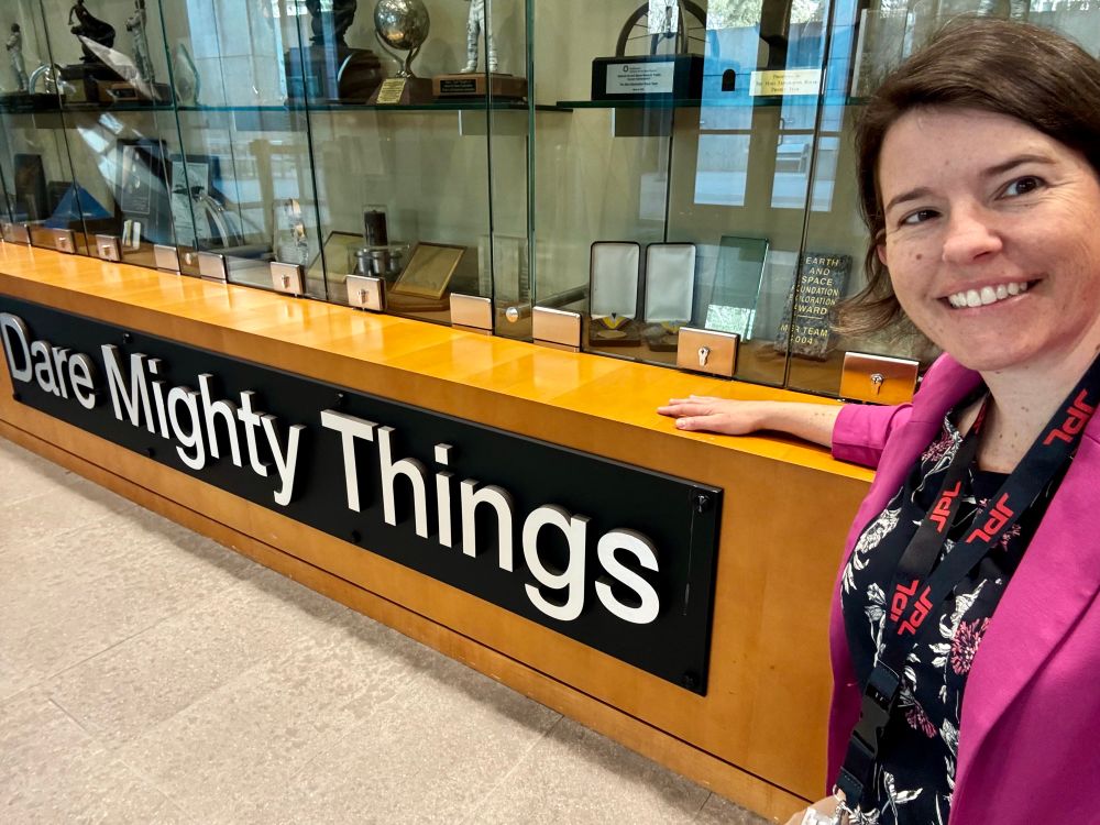 Selfie of Jessie in a pink blazer in front of a cabinet of spacecraft models labeled DARE MIGHTY THINGS. 