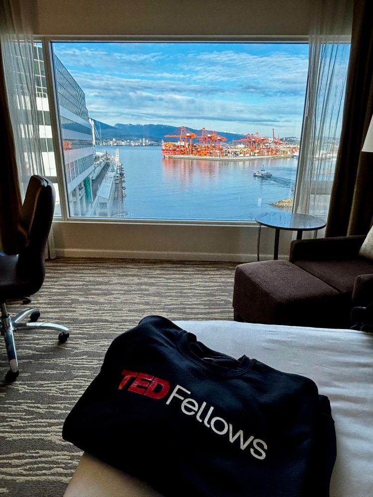 Picture of a black sweater that says TEDFellows in front of a window showing the Vancouver harbour and orange cranes