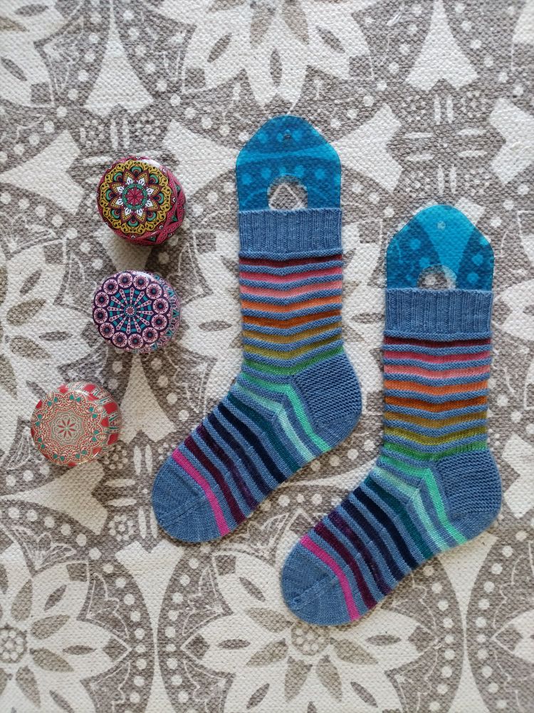 A flat lay image of a pair of hand knitted socks with stripes alongside three containers with scented candles. The socks have a denim main colour and rainbow coloured stripes. The candle containers have geometric mandala images in various colours. 