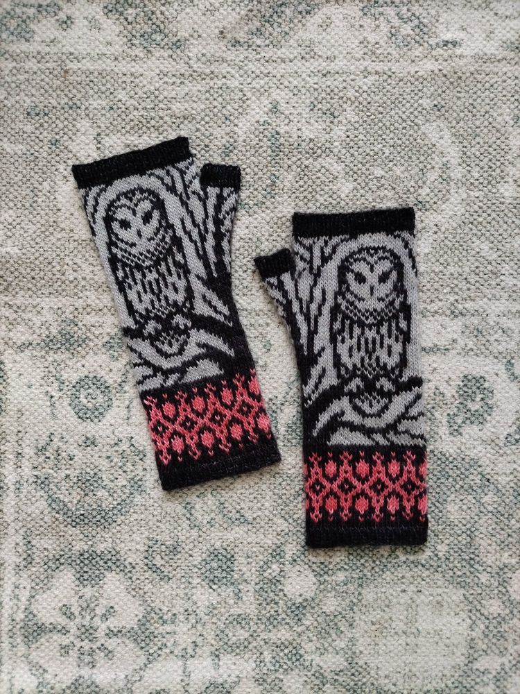 A flat lay image of a knitted colourwork mitts with owls. The mitts are black and gray with coral geometric designs at the bottom. 