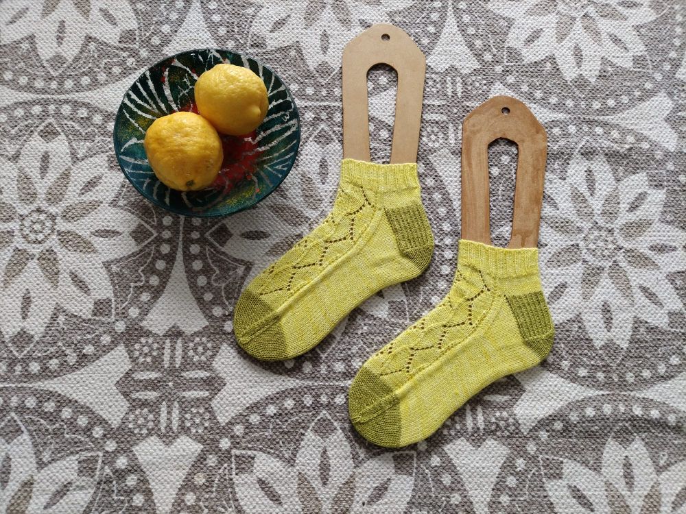 A flat lay image of a pair of hand knitted lace socks. The socks are lemon yellow with green heels and toes and are shown alongside a glass bowl with two lemons in. 