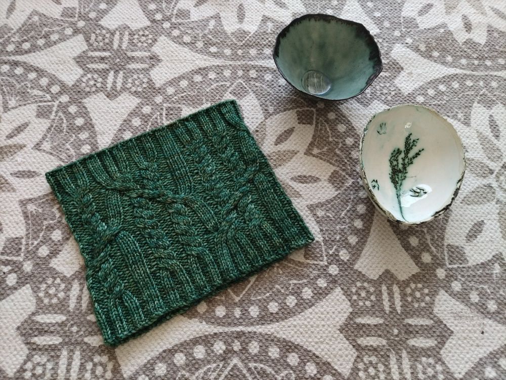 A flat lay image of a hand knitted cable cowl in green yarn alongside two ceramic dishes, one turquoise and the other white with floral imagery and birds