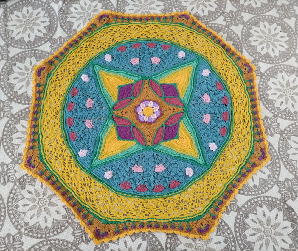 A flat lay image of a crocheted floral blanket. The blanket has a purple flower in the center surrounded by petal inlays and larger petals of green and gold. There is a large blue section with pink flowers surrounded by yellow crocheted lace. The border is ochre with purple and green flowers. 