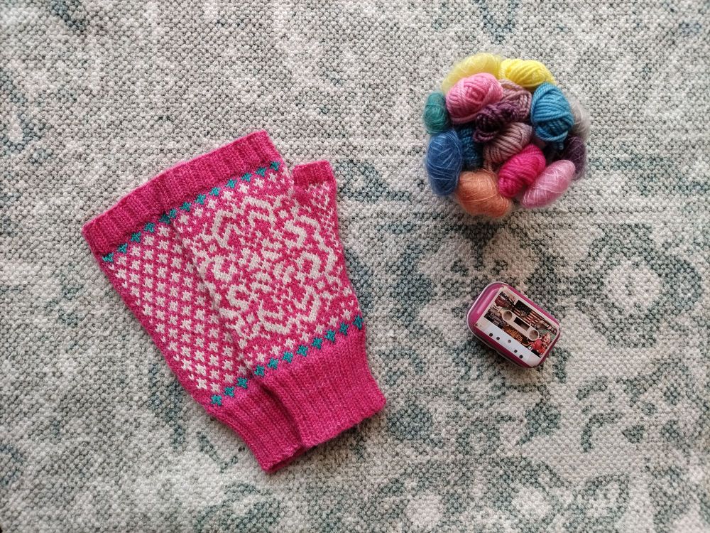 A pair of bright pink fingerless mittens with white and blue colourwork details is shown alongside a ceramic bowl filled with multicoloured yarn and a small pink tin. 