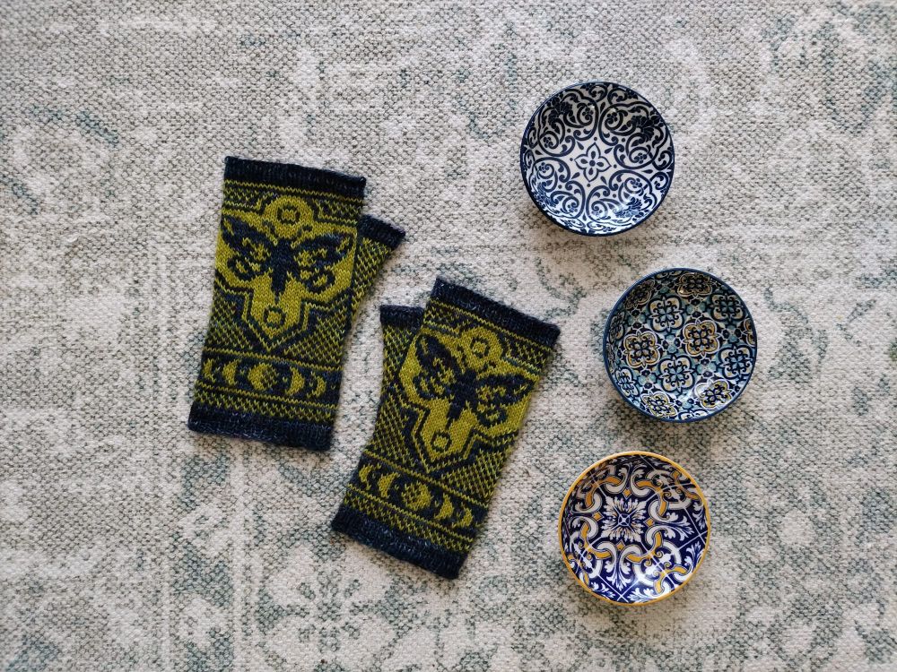 A flat lay image of a pair of hand knitted colourwork mitts, the mitts are navy and Chartreuse, and feature a underwing moth in the front, with phases of the moon. The mitts are shown alongside three small ceramic bowls with geometric designs in shades of blue, white and yellow