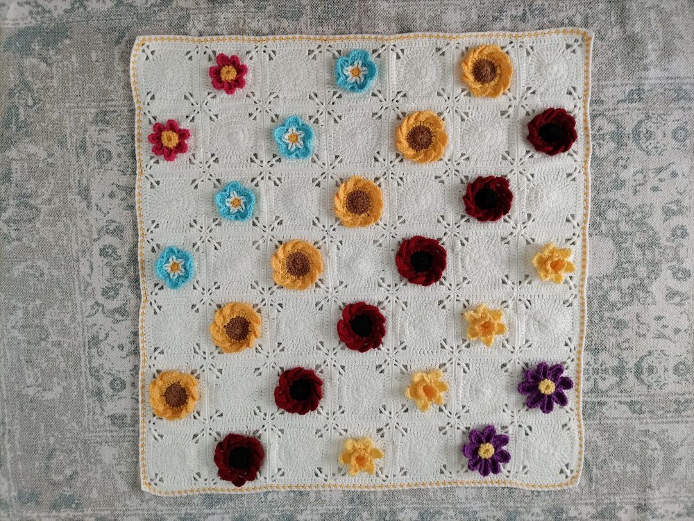 A photo of a crocheted floral blanket which has daffodils, red and yellow sunflowers, forget me nots and pink and purple cosmos with plain cream squares in between.  It has a yellow border