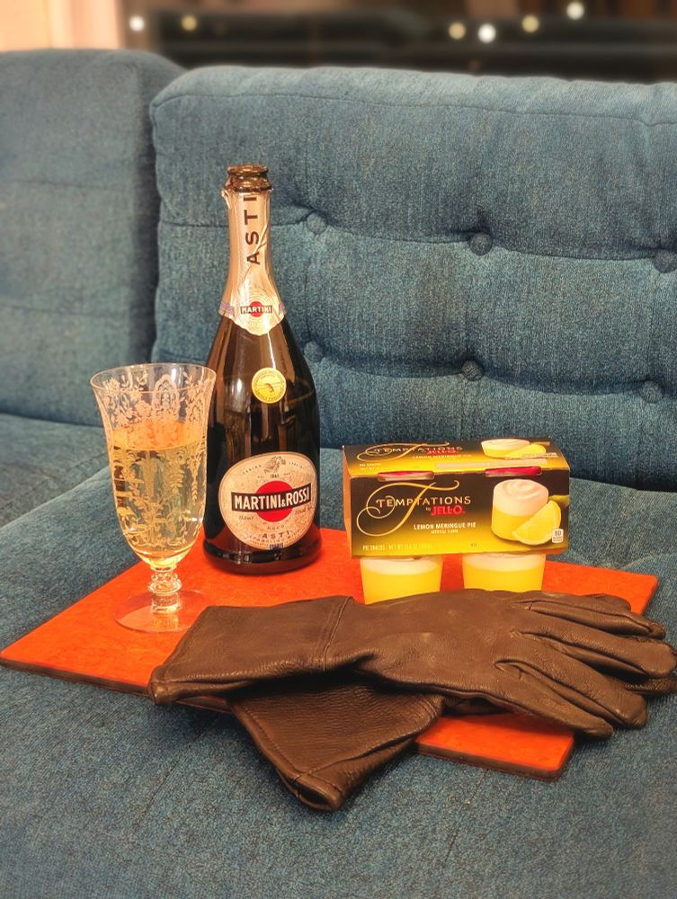 A cutting board sits on a blue couch with a bottle of Martini Rossi and Temptations Jell-O cups along with a sinister pair of black gloves