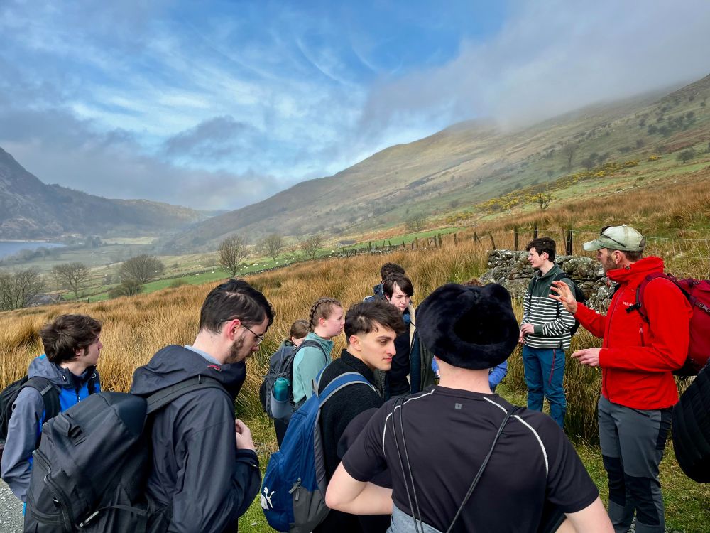 A group of young people, backs to the camera, listening to a lecturer talk about the landscape. They are stood on the side of a hill, with mountains and a large lake in the backgroud.