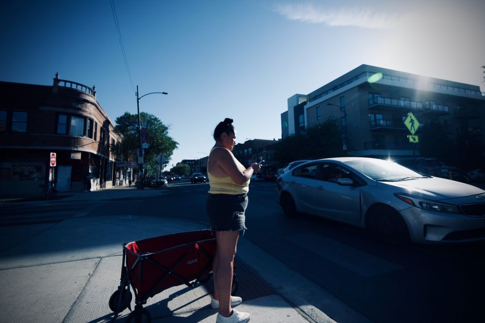 A woman pulling a red wagon behind her in an urban environment is standing at the middle of a busy intersection looking at her mobile device as cars go by