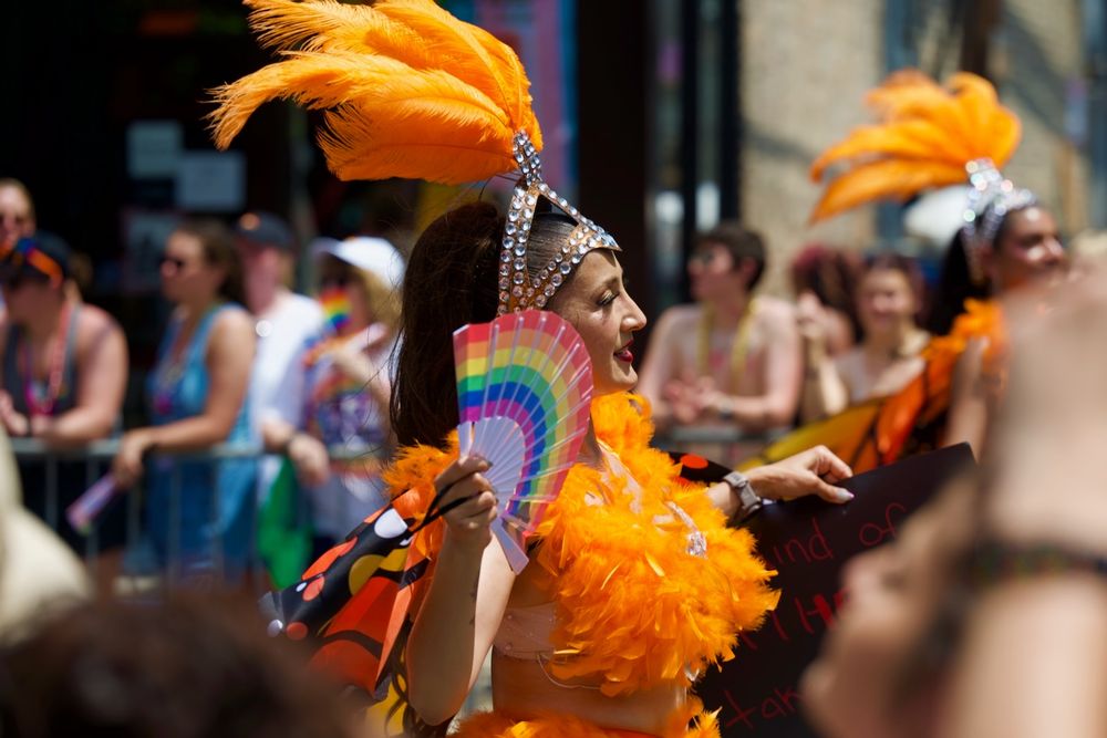 A woman in a bright orange, Mexican dance dress with a feather-laden tiara waves a rainbow fan in her face as she dances by.