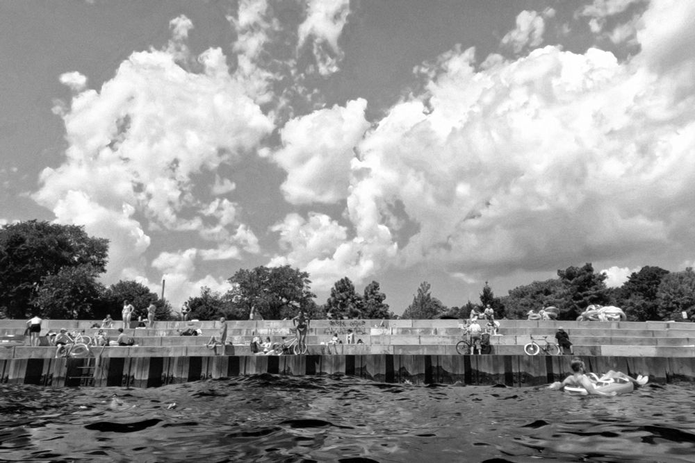 View from the water of several tiers of raised concrete walls along a waterfront with swimmers in the water, one in an inter tube. Other people in swimsuits sit along the concrete tiers enjoying bright, sunny weather. some clouds and trees are seen in the background