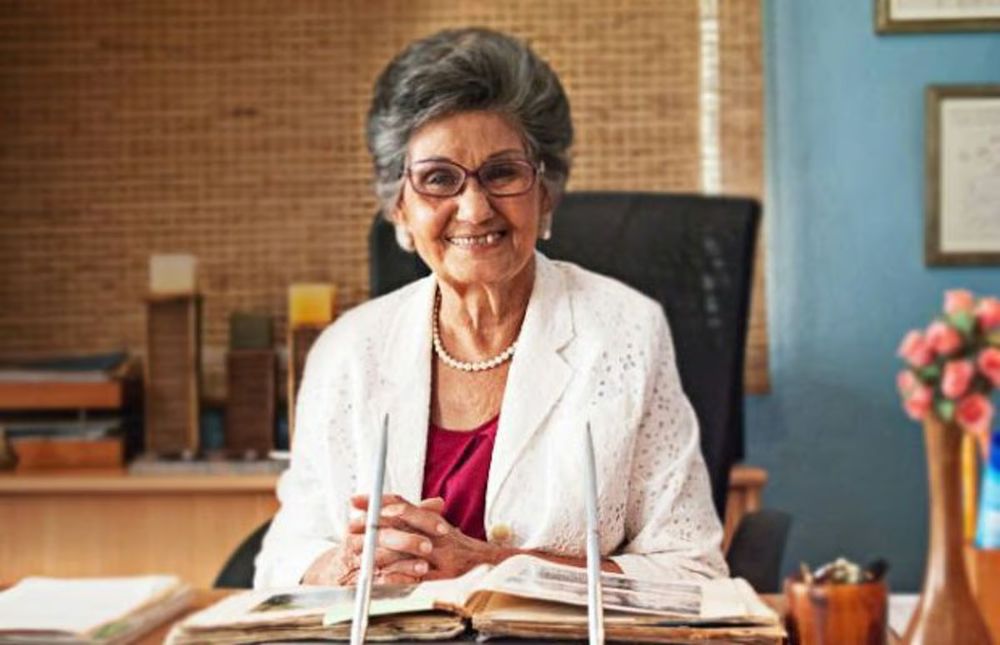  Professor Bonnelly, an older woman with light brown skin and salt-n-pepper hair. She wears a white blazer, red blouse, a strand of pearls, and glasses. She is smiling at her desk, books and papers before her.
