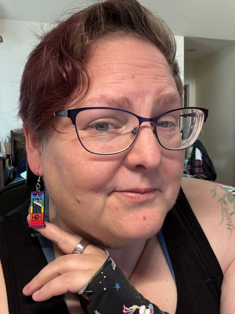 Me, a pale beige woman with short dark hair, and blue eyes, wearing purple glasses - showing off one of her new Rainbow colored Guillotine earrings. 