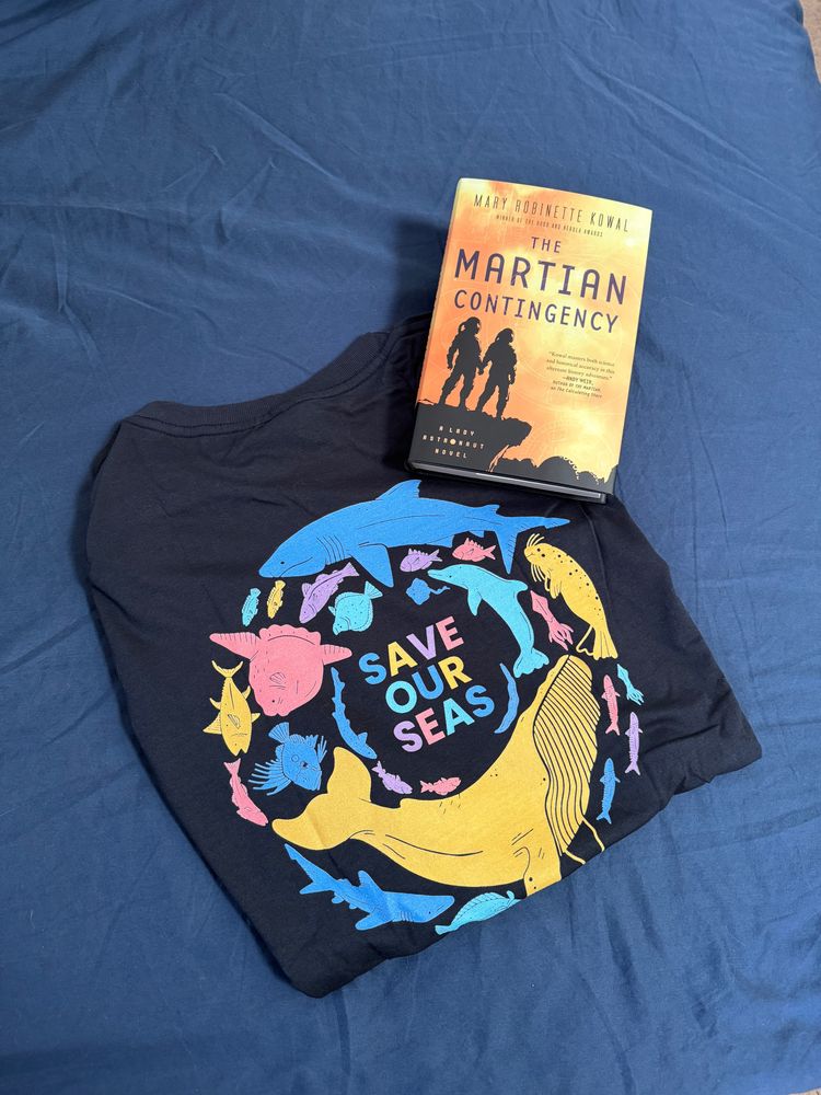 Dark blue T-shirt with a variety of large and small marine life in silhouette in blues, pinks and yellows against. They form a circle around the words SAVE OUT SEAS. At the top right of the shirt is book with an orange and yellow book cover with a two people in black on a ledge in space gear. Pale letters within the cliff read: A Lady Astronaut Novel. The book's title: the Martian Contingency is in black above the figures, with author's name above: Mary Robinette Kowal. Below the title is a quote: "Kowal masters both science and historical accuracy in this alternate history adventure." - Andy Weir Author of the Martian.