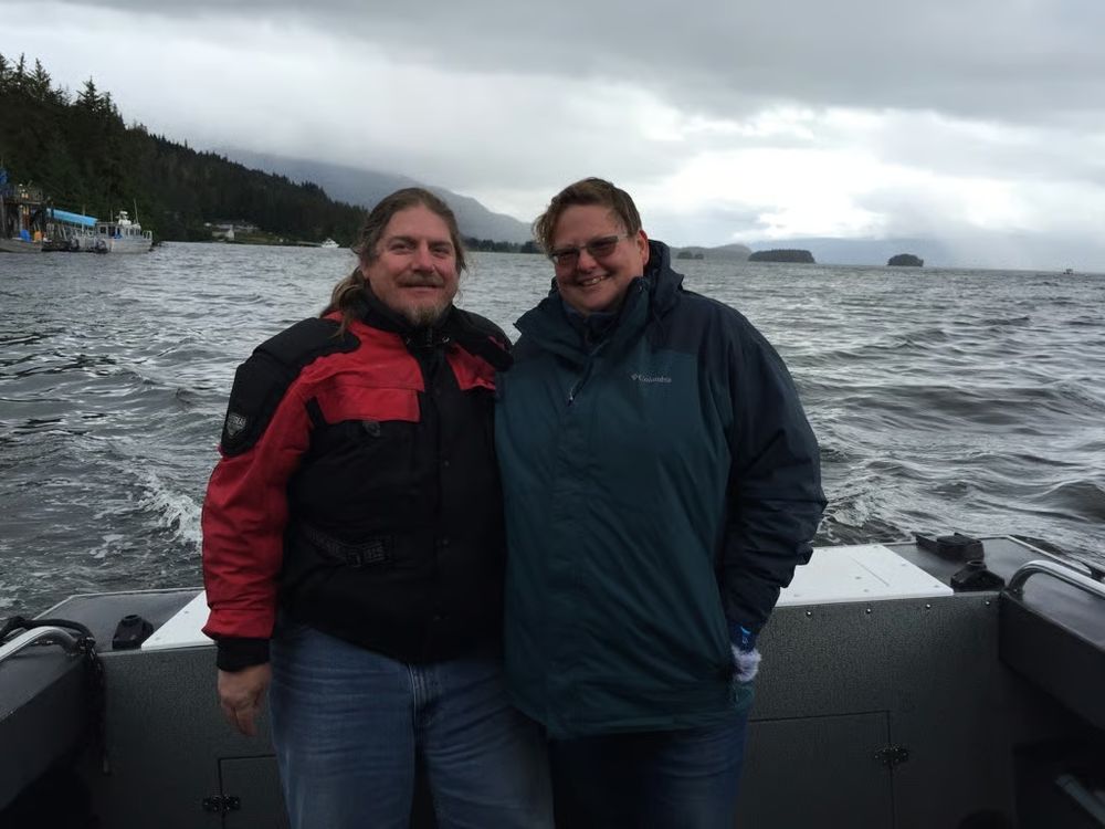 Me and the sweetie, both pink-cheeked from the wind, pose for a post-whale watch photo. Scott, on the right wears a black and red jacket, Kate on the right is in a blue windbreaker, we are standing at the back of the boat with thick fog and clouds behind us rolling toward dark tree-lined slopes leading to gray-green water.