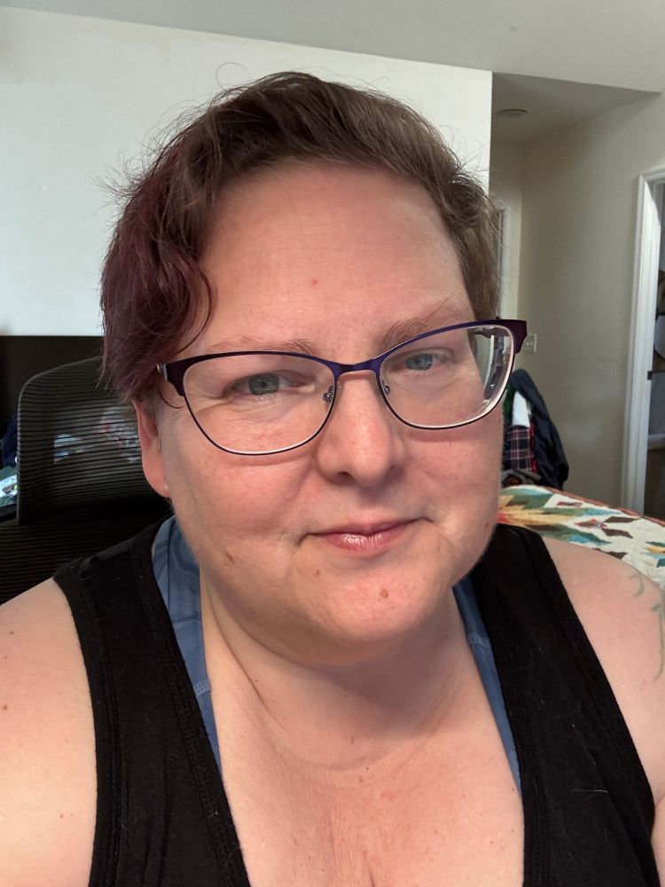 Me, a pale beige woman with short dark hair, and blue eyes, wearing purple glasses - my sort-of smile hints that I am contemplating coming out into the light of Spring. 