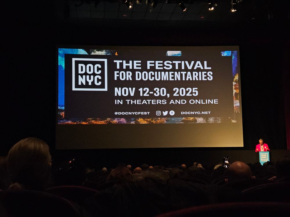 a crowded theater with an ad for doc NYC on screen. a man in a red suit stands at a podium near the bottom right of the screen.