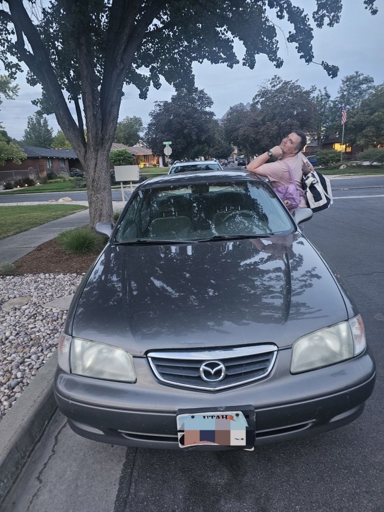 A dark grey 2002 Mazda 626 with me posing off to the side.