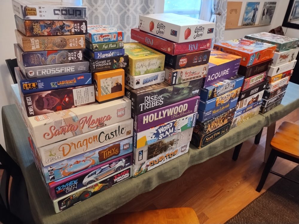 A ton of board games on a table:
A Game of Thrones: Hand of the King 
Acquire (1999 Avalon Hill Edition) 
Android: Infiltration  
Bargain Basement Bathysphere  
Bears and Bees  
Blokus 
Bravo!  
Cosmic Encounter Duel 
Coup  
Crossfire  
Cubitos
Drachenland 
Dragon Castle 
Elder Sign  
Escape from the Aliens in Outer Space  
Fuji Flush  
Hanabi  
Hollywood Blockbuster  
Jewel Heist  
Joyride: Duel - Next Gen  &  Joyride: Survival of the Fastest  
La Granja: No Siesta  
Labyrinth the Card Game  
Letter Jam  
Lost Ruins of Arnak  (includes the Missing Expedition and Expedition Leaders expansions 
Master Labyrinth 
Mining Colony  
Monster Crunch! The Breakfast Battle Game  
O Zoo le Mio 
Quoridor  
Raj  
Red Cathedral  
Reign of Dragoness 
Rise of Tribes 
Sanctum
Santa Monica  
Say Anything
Scotland Yard
Scrabble 
Skull King  
Stinker
Sword and Skull 
Team Play  
Tiki Topple  
Tiny Towns  
Valletta  
Word Domination
Word Slam 
World Wonders
