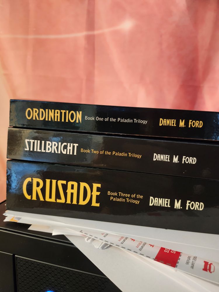 A stack of books, the Paladin Trilogy by Daniel M Ford. Top to bottom they are titled Ordination, Stillbright, and Crusade. 