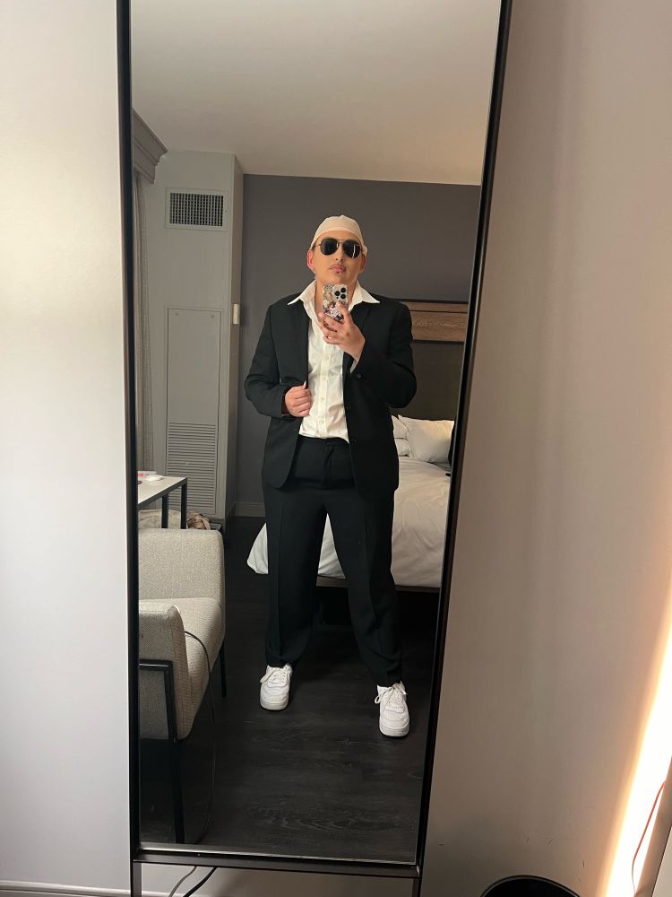 Celeste is dressed as Pitbull, aka Mr. Worldwide. She’s wearing a bald cap, black aviator sunglasses, white button down shirt, black blazer, black pants and white sneakers. She’s posing in front of a hotel mirror. 