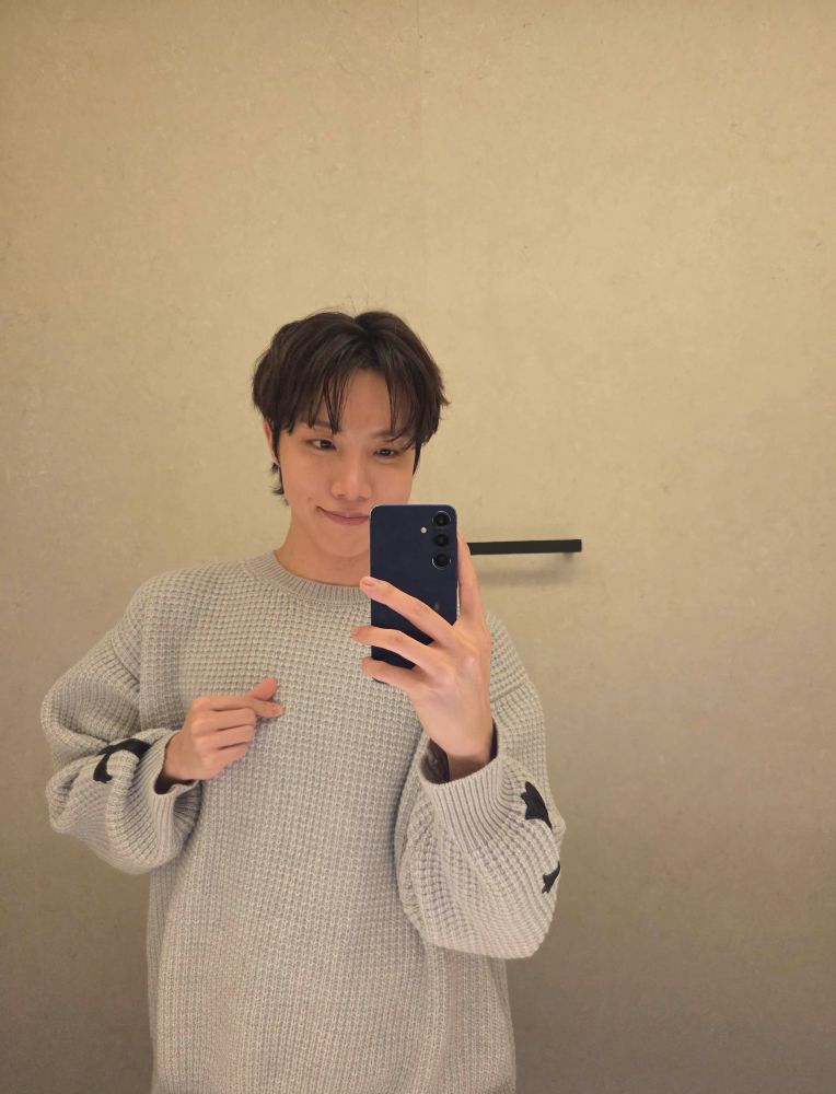 a very soft beige photo of hobi looking like I just knocked him up and he's very excited about it. he's giving finger hearts and is wearing a huge oversized beige sweater and his little dimples are showing 