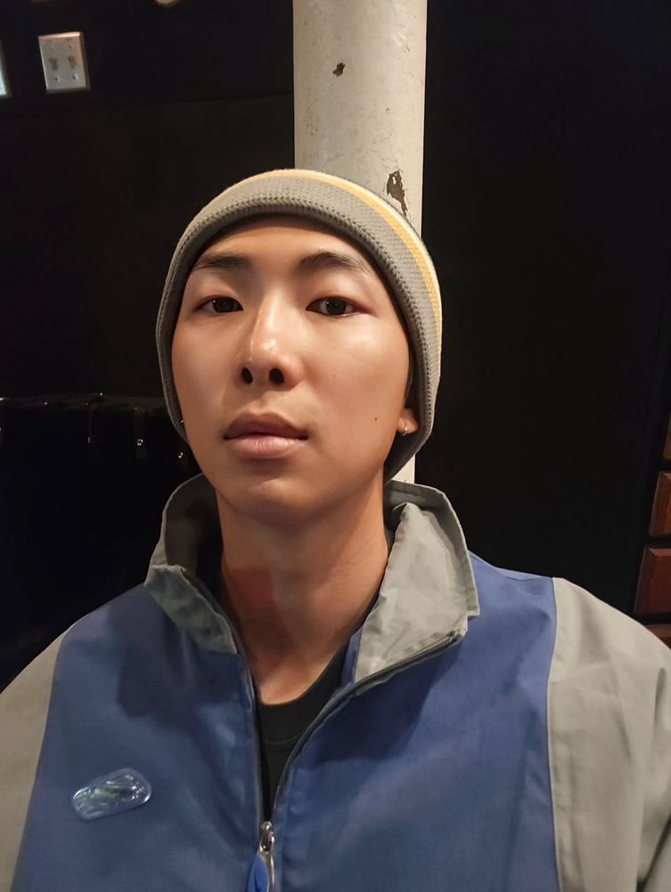 Namjoon in a windbreaker and beanie, staring directly at the camera without smiling. It's same face and angle San Yawn uses in a lot of his selfies, actually. Joon is tan and looks beautiful and you can see how gorgeous his features are, especially his eyes and his full lips. 